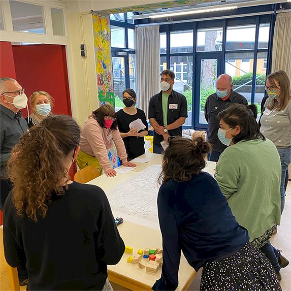 Um einen Tisch herum versammelt, stehen insgesamt zehn Personen. Auf dem Tisch ist eine Stadtkarte ausgebreitet.