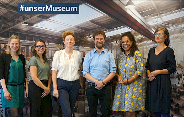 There are six people shown in the group photo. They are standing in front of a photo wall. The photo wall shows a section of the design of the future migration museum. On the top left of the wall is the writing #unserMuseum.