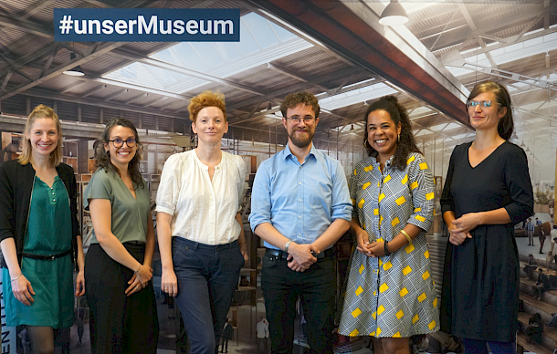 Auf dem Gruppenfoto sind sechs Personen abgebildet. Sie stehen vor einer Fotowand. Auf der Fotowand sind ist der Ausschnitt eines Entwurfs des zukünftigen Migrationsmuseums zu sehen. Auf der Wand oben links steht #unserMuseum.