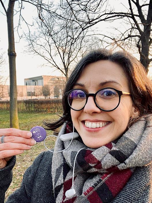 Sandra Vacca lächelt mit dem den Button von DOMiDLabs in die Kamera. Im Hintergrund das Kanzleramt in Berlin.